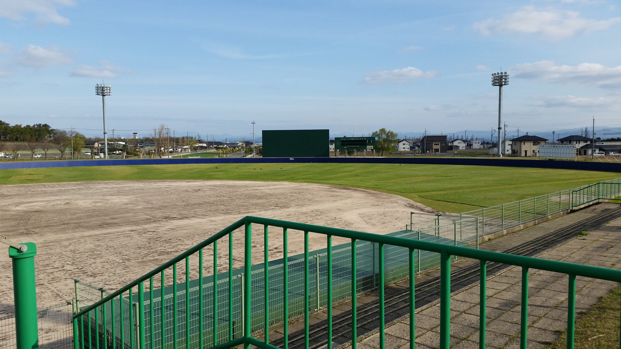 Empty dirt and grass sports field viewed from stands, green railings, light towers, houses in distance under blue sky