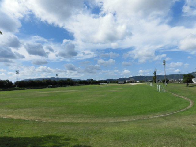 Wide grassy sports field with soccer goals, distant tree line and mountains, large cloudy blue sky