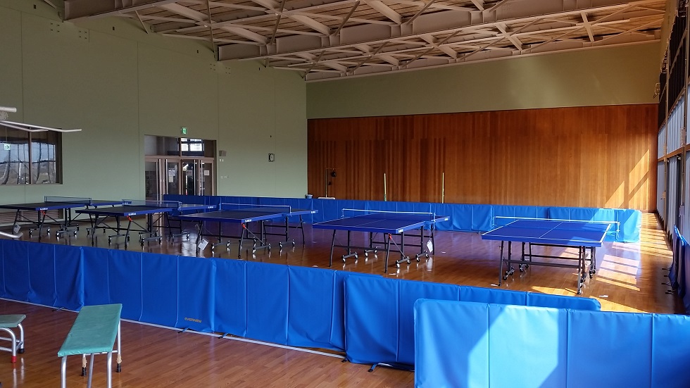 Indoor gym with multiple blue table tennis tables on wooden flooring and blue padded wall barriers