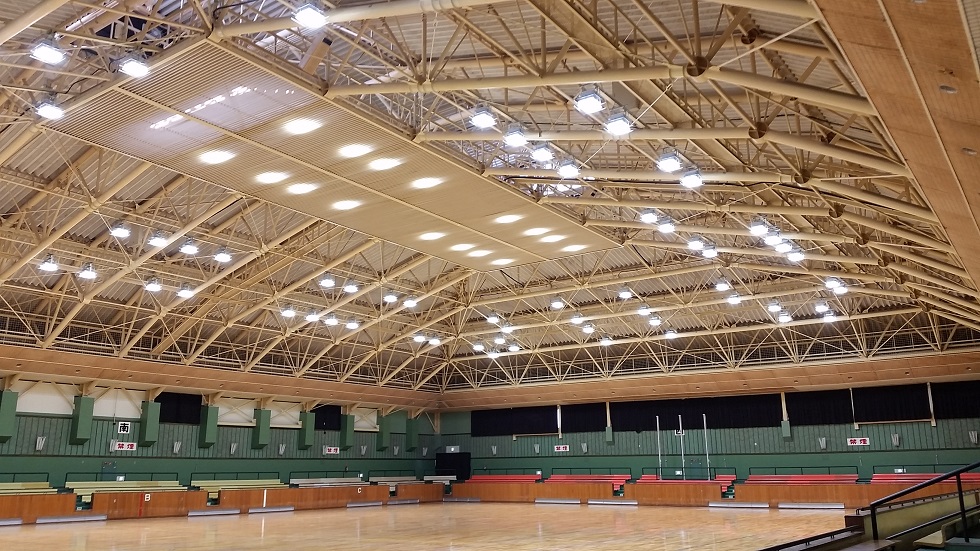 Large indoor gymnasium with high ceiling, exposed metal trusses, wooden floor, and rows of bleacher seating, bright and spacious